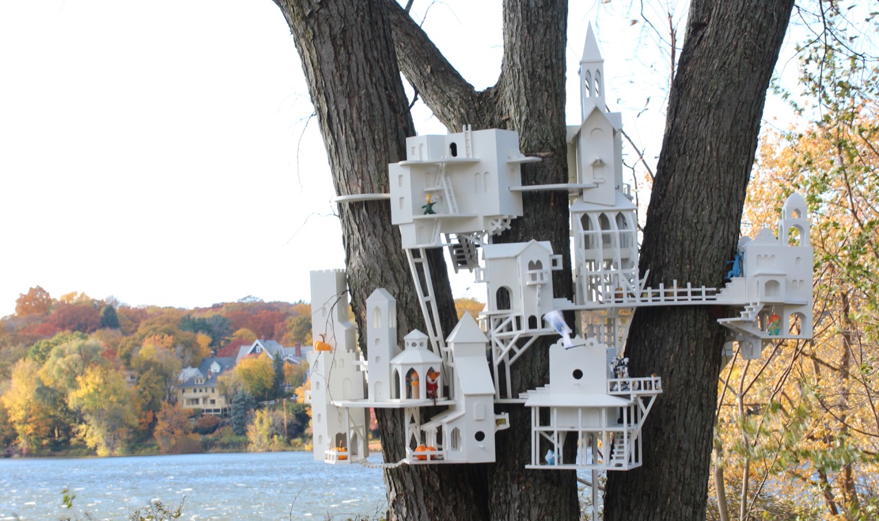 in the foreground, a complex miniature treehouse with various styles of architecture; in the background, a lake and the opposite shore beyond, with fall foliage and houses that are on the same scale as the treehouse (from this distance).