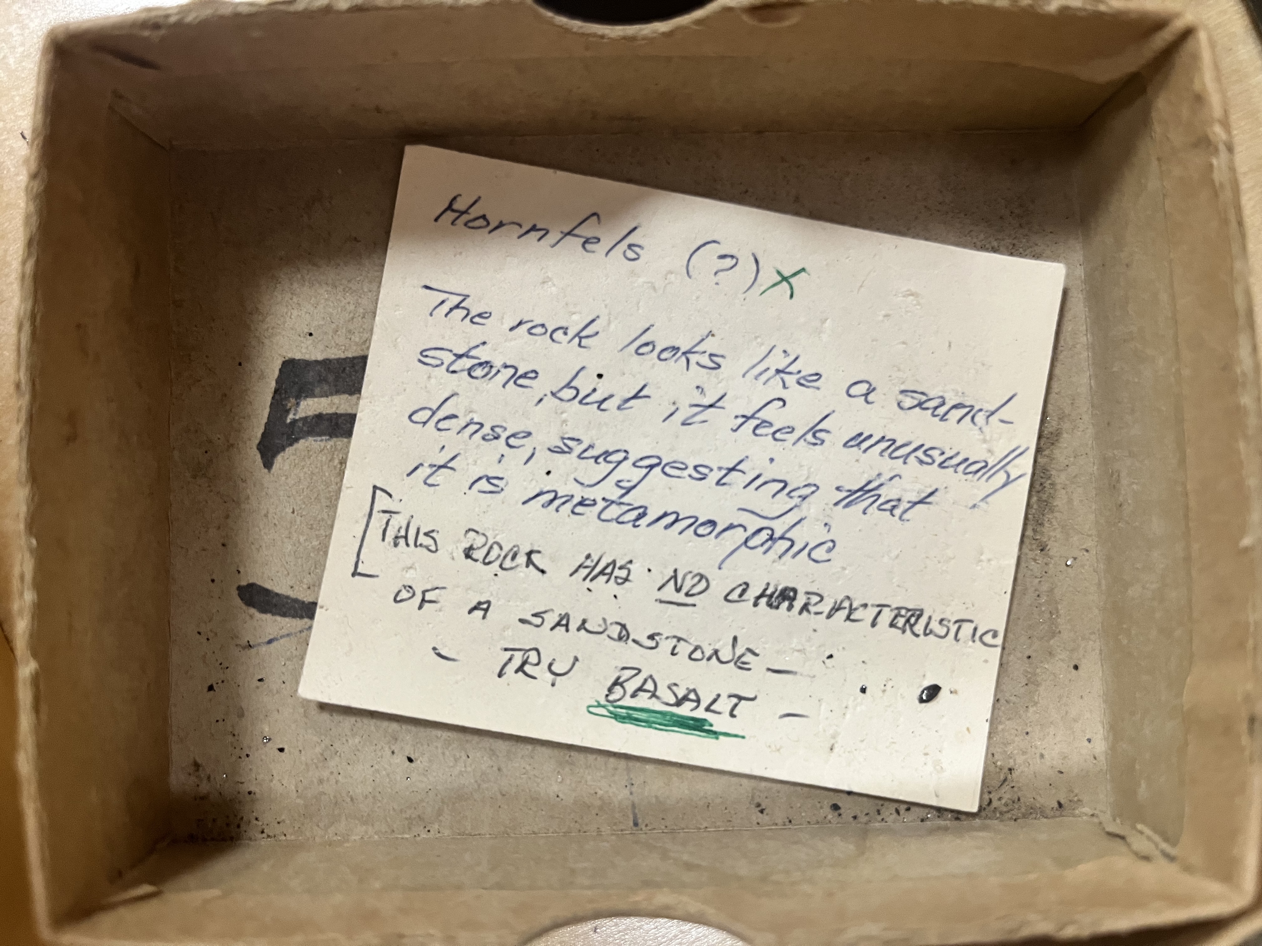 a somewhat yellowed piece of paper in a cardboard box. it reads "Hornfels (?) X / The rock looks like a sandstone, but it feels unusually dense, suggesting that it is metamorphic". below this a note in different handwriting reads "THIS ROCK HAS NO (underlined) CHARACTERISTICS OF A SANDSTONE -- TRY BASALT (heavily underlined)"