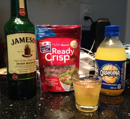A bottle of Jameson whisky, A package of Ready Crisp bacon bits and a bottle of Orangina. In front of those is a shotglass with the mixed drink in it.