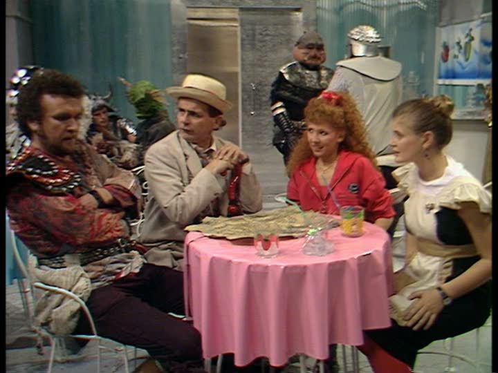 Screencap from Dragonfire: in a 1950s-style soda shop, four people sit around a table: from left to right, Sabalom Glitz (a space pirate with a short beard), the Doctor, Mel, and Ace (wearing a waitress's uniform).