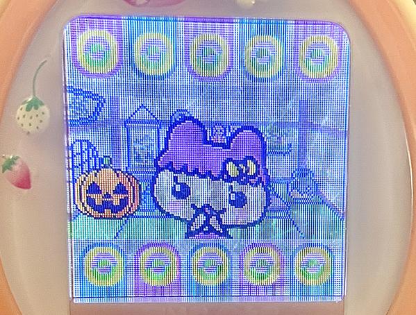 close up photograph of a tamagotchi plus color device. chamametchi is on the screen, close to the viewer, and posing cutely. a festive jack-o-lantern is in the room with her.