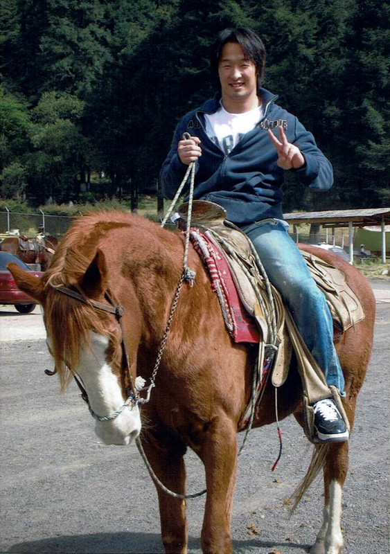 Photo of Naomichi Marufuji riding a horse. He is smiling and giving a peace sign to the camera.