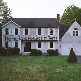 A large white house on a misty day, with the title of the album spray painted on the siding