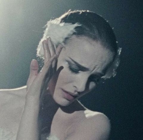 A still from the Black Swan movie. Nina, the main character portrayed by Natalie Portman, is dressed in a white swan costume, complete with ghostly makeup and a swan headpiece. She is looking down with scrunched eyebrows and a face that expresses pain of some sorts. Her left hand is facing outward as it drapes on her cheek.
