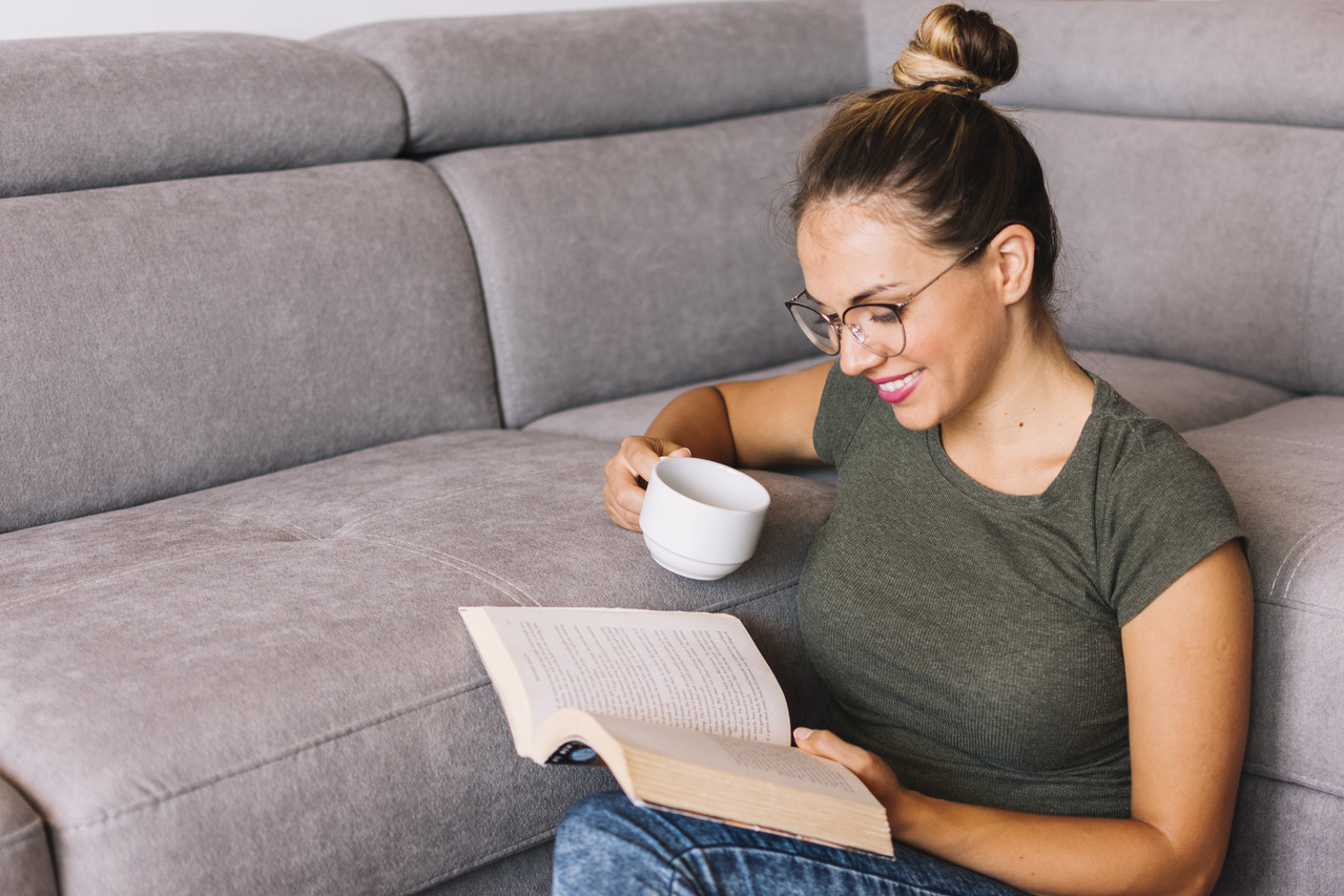 Smiling young woman reading a book and holding a cup of coffee near a sofa