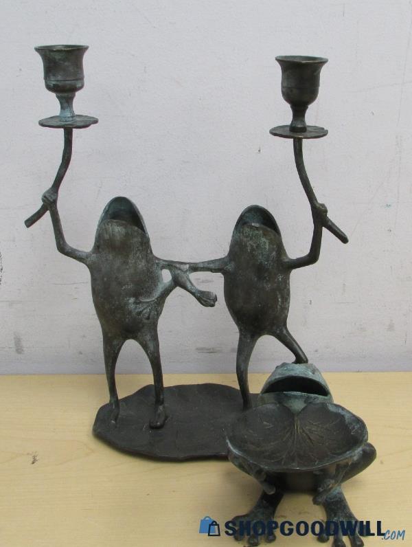 a product photo of a metal candelabra shaped like two frogs holding up flowers while standing arm in arm and shouting at the sky. in front of the candelabra is a matching soap dish, shaped like a frog offering a lily pad.