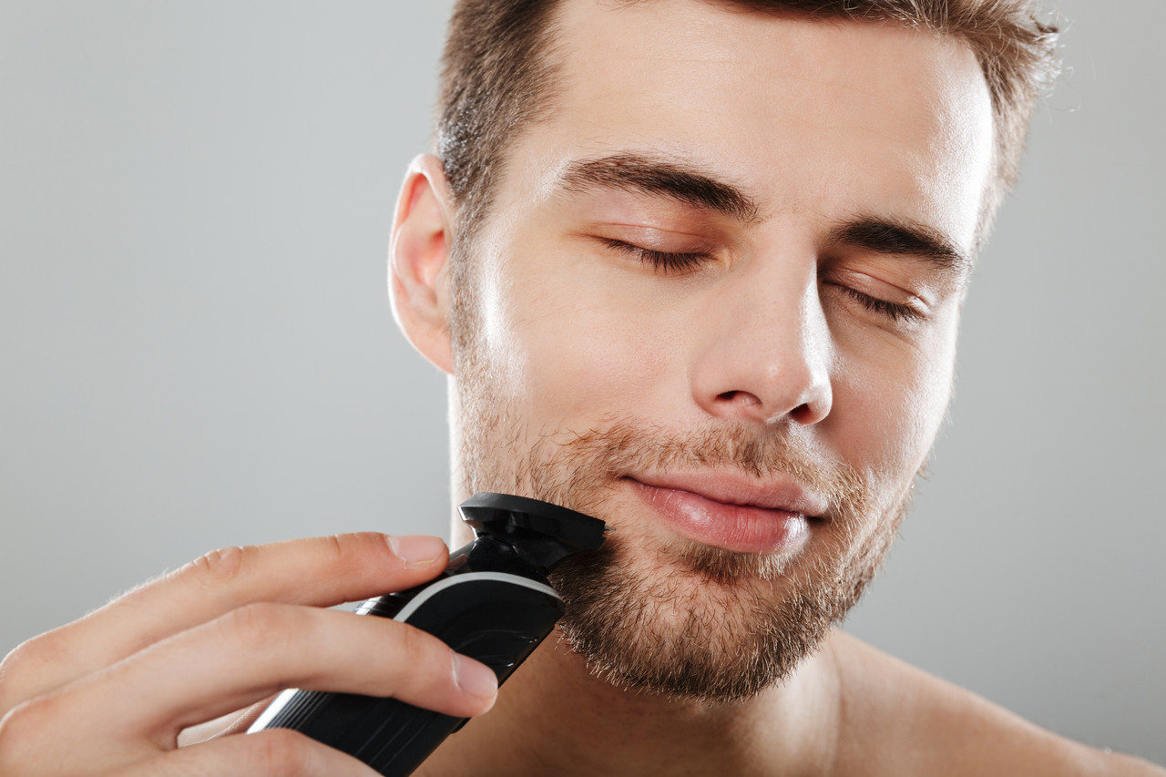 headshot caucasian pleased guy 30s being undressed bathroom while shaving face with trimmer grey wal