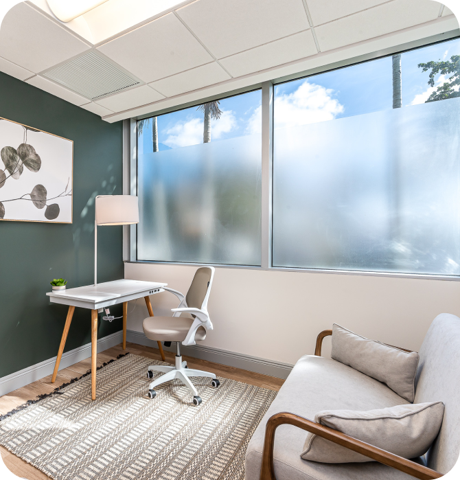 Bright corner office with large windows offering a view of palm trees outside. The space has two desks equipped with laptops, creating an inviting and productive environment.