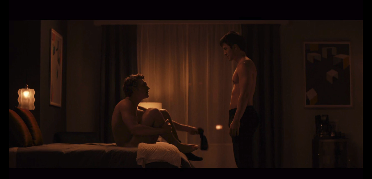 ilya is sitting on the bed naked, taking his socks off. shane stands at the end of the bed, shirtless and in black pants. 