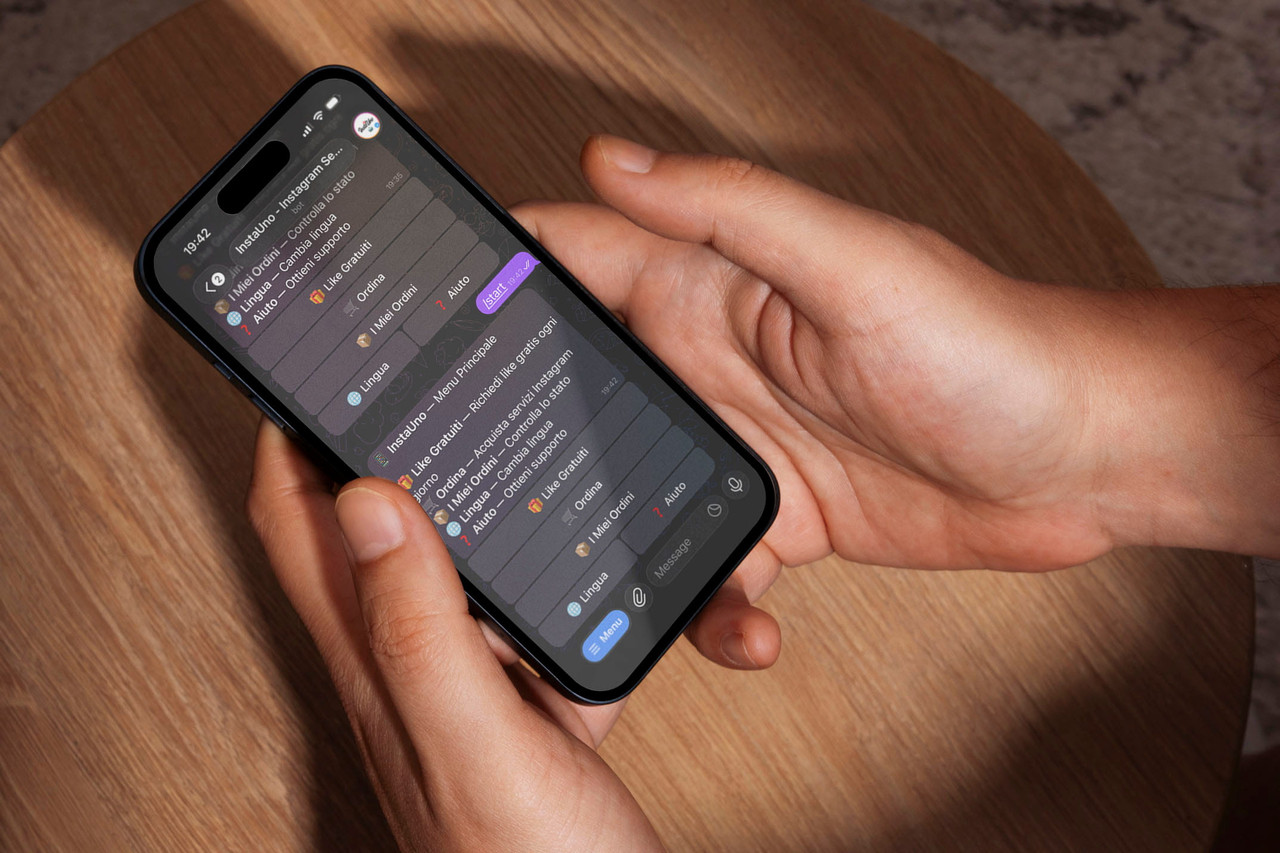 Mani che tengono uno smartphone mostrando la chat del bot Telegram InstaUno con le opzioni per i servizi Instagram, sopra un tavolo di legno.