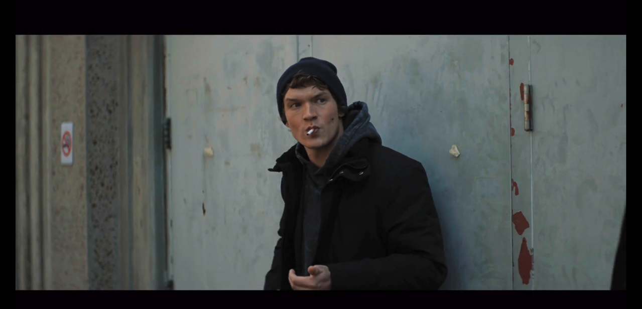 ilya stands leaning against an exterior wall with a cigarette in his mouth. he's wearing a black beanie and a dark gray sweatshirt under a black button-up coat.