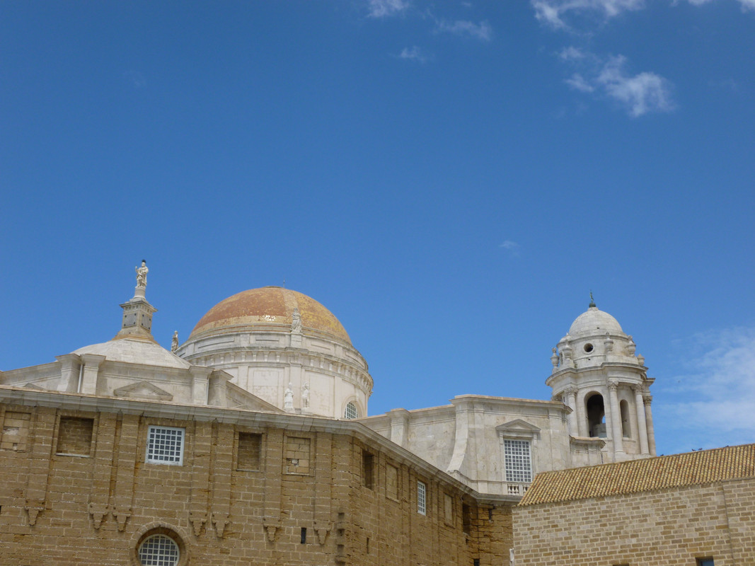 Photo of the back of the old cathedral.  In the centre is a white tower with an orange dome, which belongs to the new cathedral.  On the left is a more pointy white tower, with a figure on the roof, belonging to the old cathedral.  Not sure who the figure is supposed to be but it looks like a Roman soldier to me.  On the right is another white tower, this time with a white dome, also belonging to the new cathedral.  In the front are two brown walls.  The one on the left looks like stone, the one on the right like brick. I