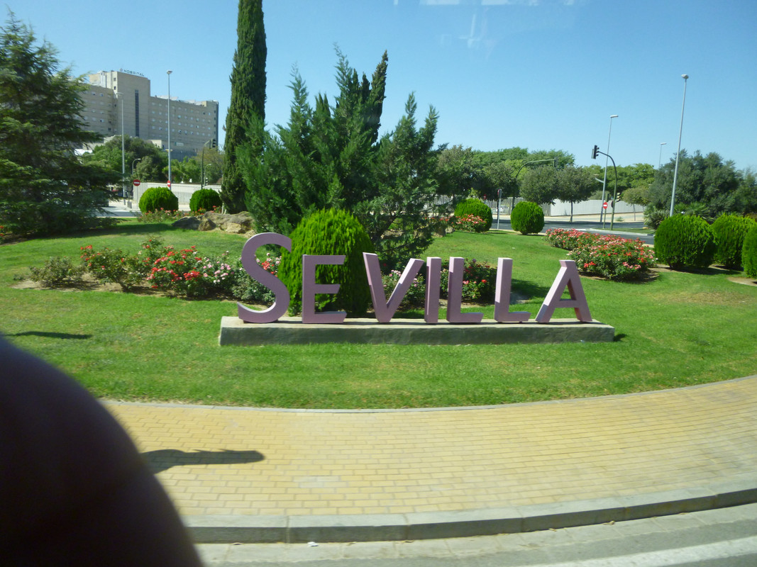 Pinky white sign saying SEVILLE in capital letters at a roundabout with grass, fir trees and some flower bushes