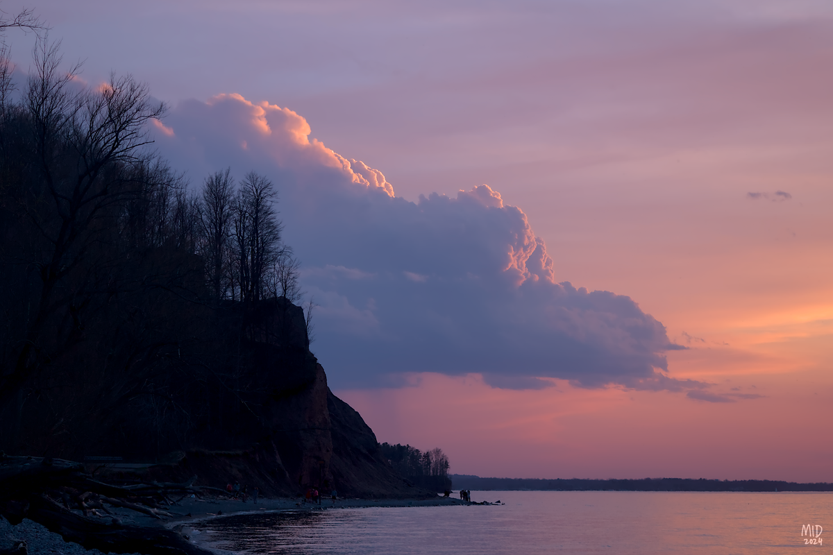 Picture of a very large cumulus cloud over a lake at sunset, partially blocked by the silhoutte of a forested hill on the left. The sunset paints the lake, sky, and top of the cloud in soft blues, purples, and oranges.