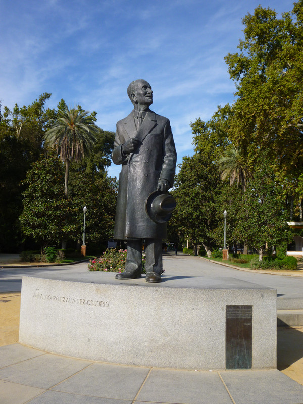 Statue of Aníbal González who designed the Plaza de España.  The statue is of a balding elderly man, wearing a double breasted long coat, and shirt with a tie and smart trousers, holding a Homburg hat.  He is looking to his left, at the Plaza.