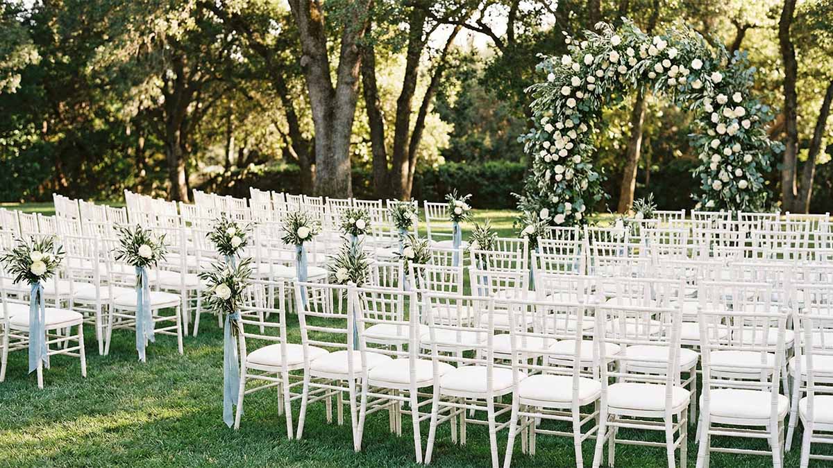 Белые деревянные стулья Кьявари, расставленные ровными рядами на зеленом газоне для выездной регистрации.