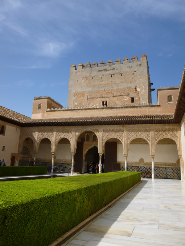 Very mudejar-style building.  There is a very plain tower in the background, the Comares tower.  There are arches which separate the walkway around the courtyard from the courtyard.  The stonework is intricately decorated.  There are tiles on the wall to about midway, and the rest is white painted.  There is a row of myrtle bushes in the foreground.