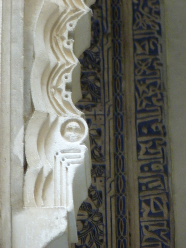 At the front of the photo is a carved arch, all in very white stone.  In the back, another carved wall, in very white stone with blue inlays.  In the front carved arch there is a shape that looks like a human skill.  The three right angles underneath it make it look like a skull attached to a rib cage