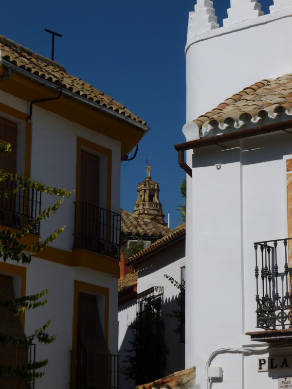 Two white-walled houses, with a few tree branches coming in the left hand side of the image. The house on the left has yellow window frames. Between the two houses you can see the top of the tower of the Mezquita.