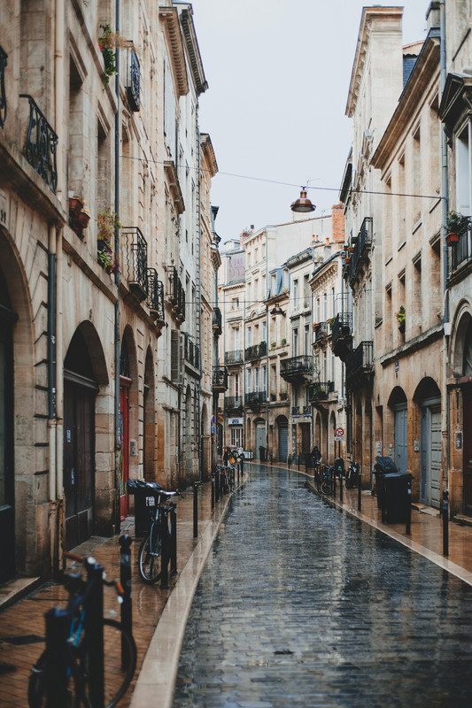 A narrow cobblestone street in a European city, lined with historic beige stone buildings featuring wrought-iron balconies and arched doorways, with bicycles parked along the sidewalk on a rainy day.