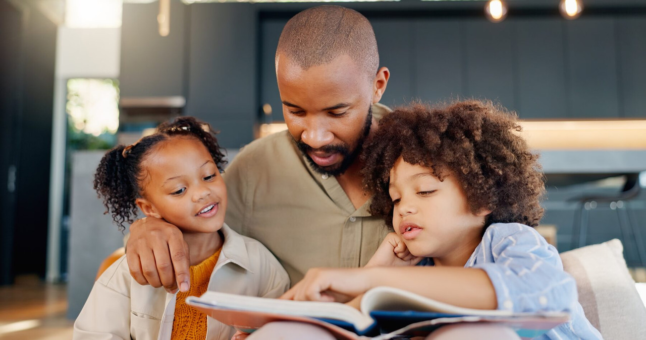 A father reads a picture book with his two children curled up beside him.
