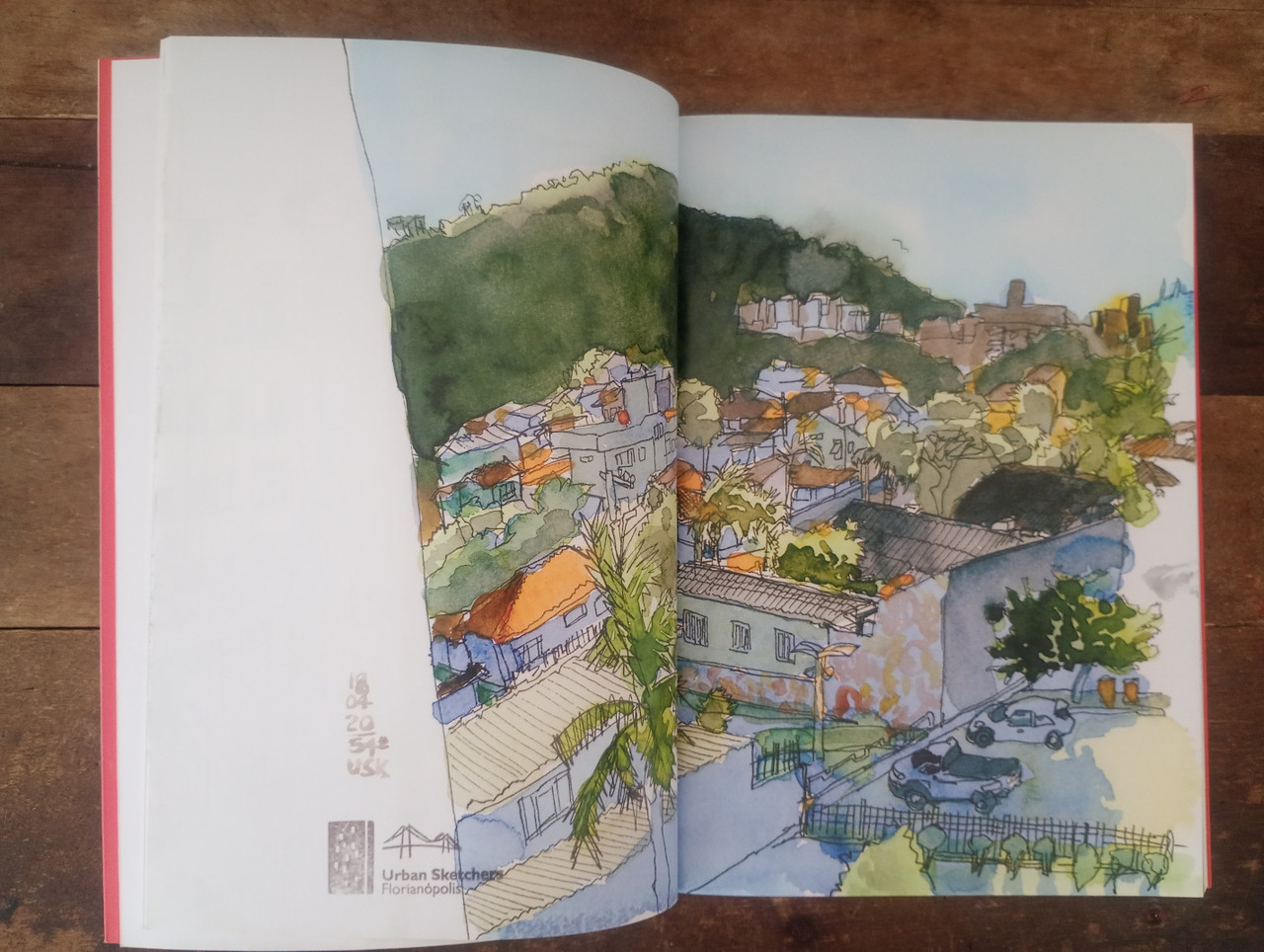 Livro aberto ao meio, em que se vê outro desenho em aquarela mostrando uma visão panorâmica da cidade de Florianópolis. A ilustração toma duas páginas.