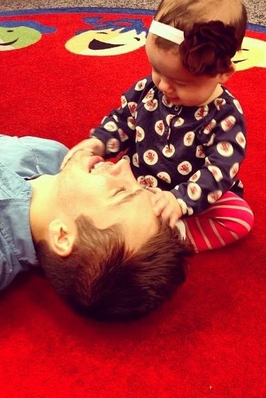 [A photo of Tyler in his early 20s with fluffy hair and a blue button up. He is laid on his back on a red playmat whilst a 8 month old baby is sat next to his head. The baby girl is trying to grab his hair with one hand whilst the other is on Tyler's chin. Tyler is smiling widely at the baby.] 