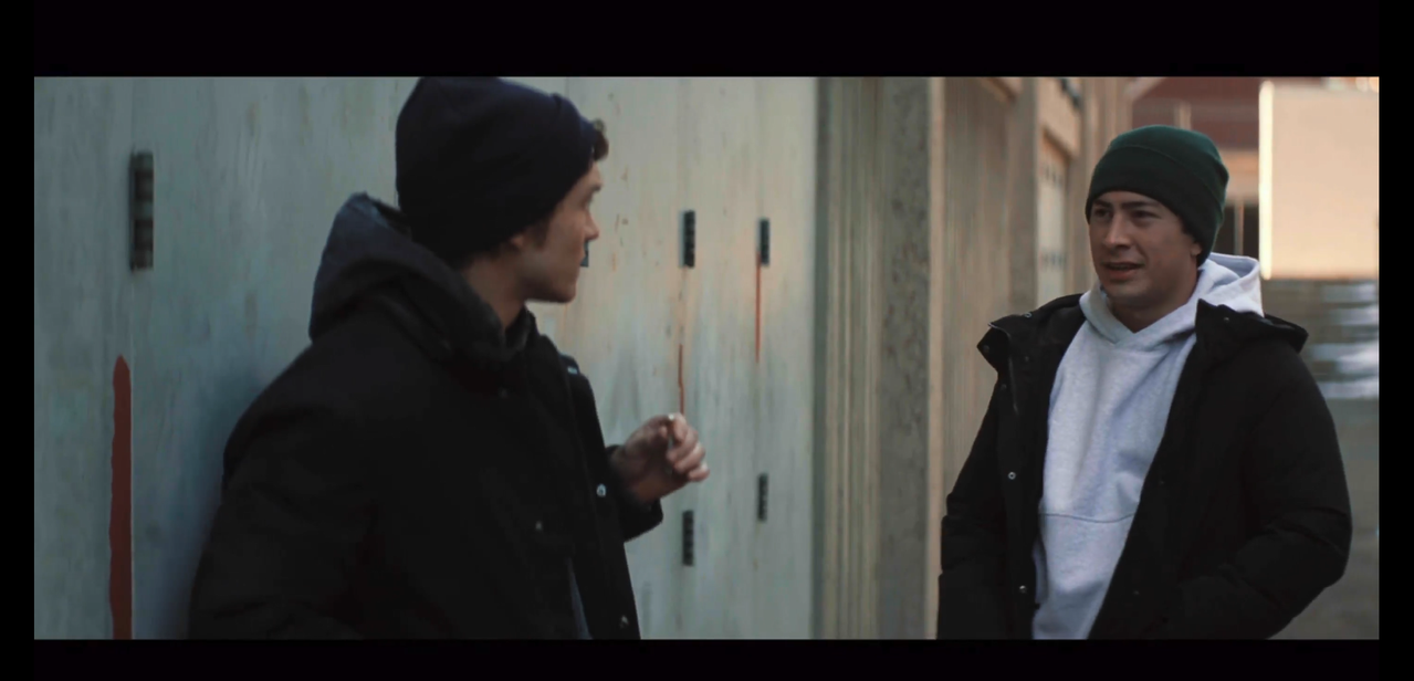 ilya stands against an exterior wall with a cigarette in his hand, looking at shane entering the frame. ilya is on the left and shane is on the right. ilya is wearing a black beanie, dark gray sweatshirt, and black button-up coat. shane is wearing a dark green beanie, white sweatshirt, and black button-up coat. ilya's on the left and shane's on the right. 
