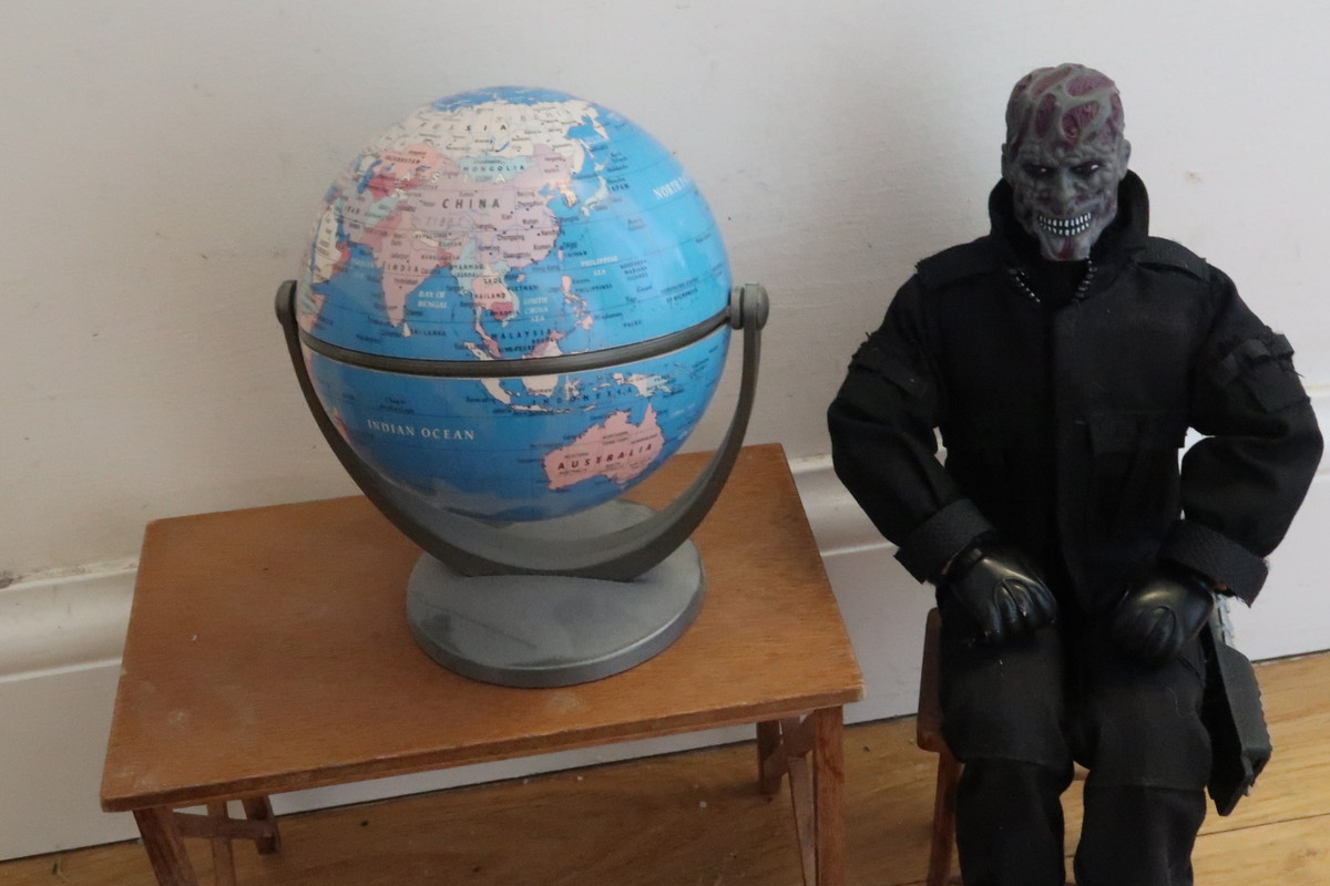 No Face sitting on a stool beside a table and a globe of the world.  37-C7-B978-2793-4774-922-D-78-C8-E86-AD76-D
