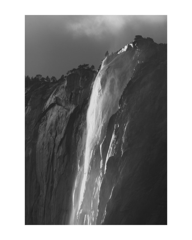 A black and white photograph of Horsetail Falls in Yosemite National Park, California, USA. The rock cliffs are dark and reflect the light where they have been made wet by the fall's spray. The sky is a cloudy grey.