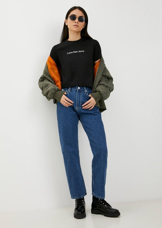 Модель в очках: чёрная футболка с логотипом Calvin Klein Jeans, оливковый бомбер с оранжевой подкладкой на плечах, синие прямые джинсы с высокой посадкой и чёрные массивные ботинки на светлом фоне.