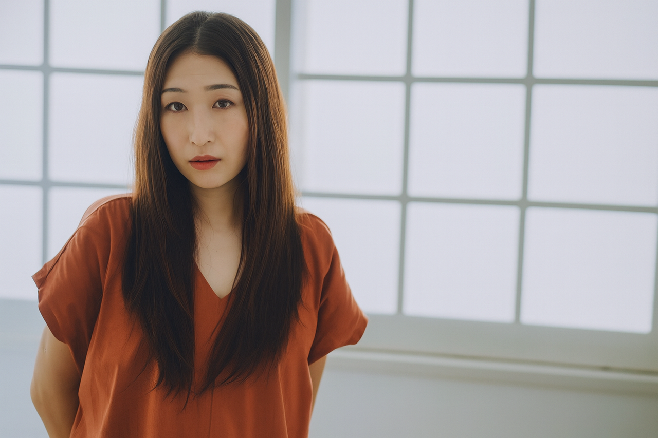 レンガ色のブラウスを着た長い黒髪の女性が 窓辺でニュートラルな表情でカメラを見つめる