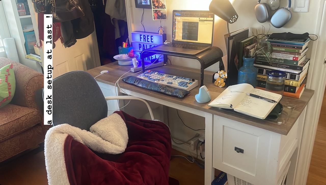 a photo of a desk with a computer riser on it, a computer on top of that, and an ergonomic keyboard & mouse and a wrist rest in front of it. on the right side of the desk, a journal is open and a stack of books sits. on the other, a little sign that says 'free palestine!' and some photos are up. a chair in front has a heated blanket draped over it.