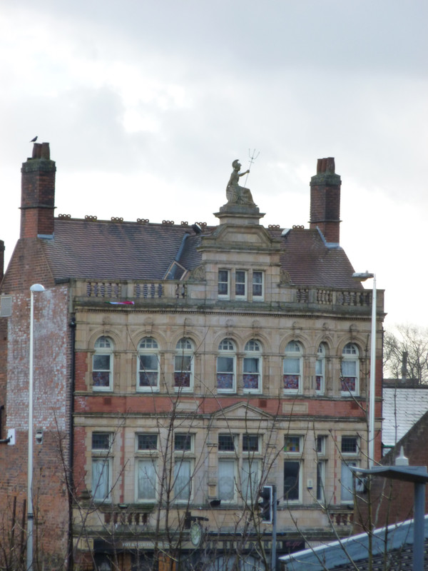 Top two floors of a disused Victorian pub.  The floor at the bottom of the image has 6 narrow rectangular windows, three either side of a larger bay window.  The floor above has 8 windows across the same span.  The windows are rectangular but the top side has been replaced by a semi-circle.  Then comes the roof and on top of the roof is a statue of what I think is Britannia because she is a woman in a chariot holding a trident.