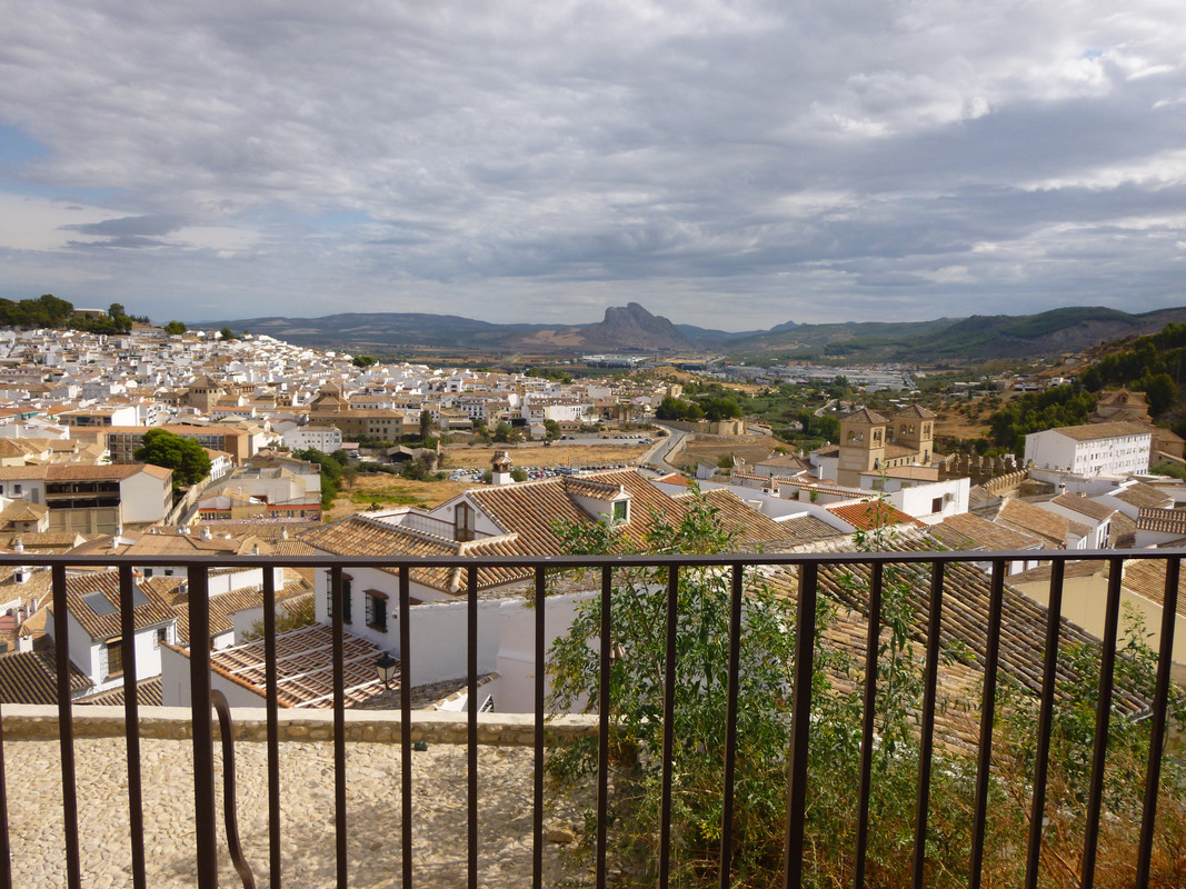 A panorama of white walled houses with orange tiled roofs.  At the front are the black metal rails of a balcony.  In the distance is a mountain that looks like the face of a giant lying down.