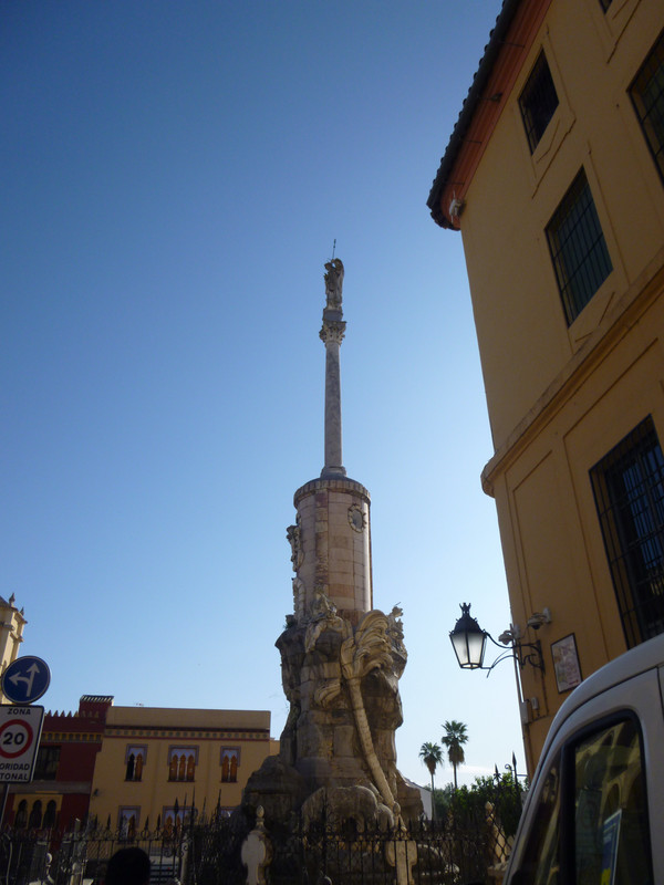 A white stone statue of St. Raphael on top of a pillar.  On the near side of a pillar a monster that looks like a dragon is climbing up.