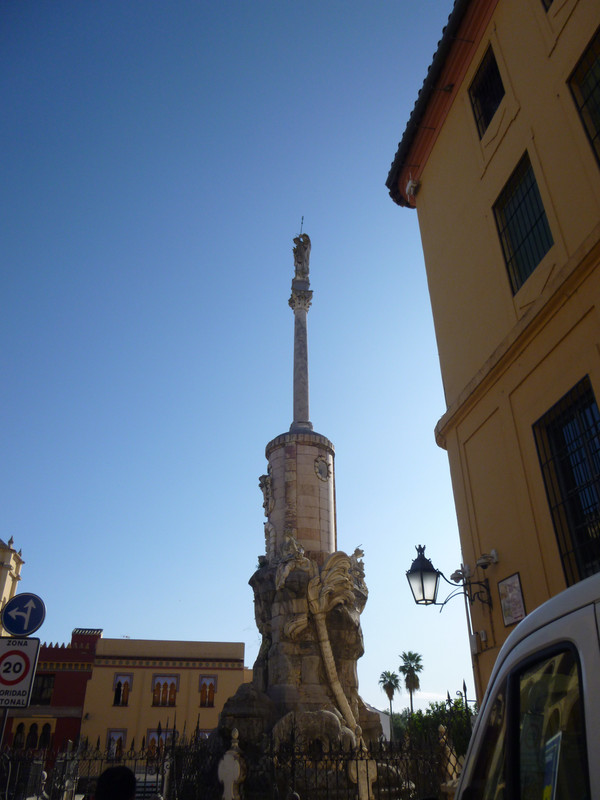 A white stone statue of St. Raphael on top of a pillar. On the near side of a pillar a monster that looks like a dragon is climbing up.