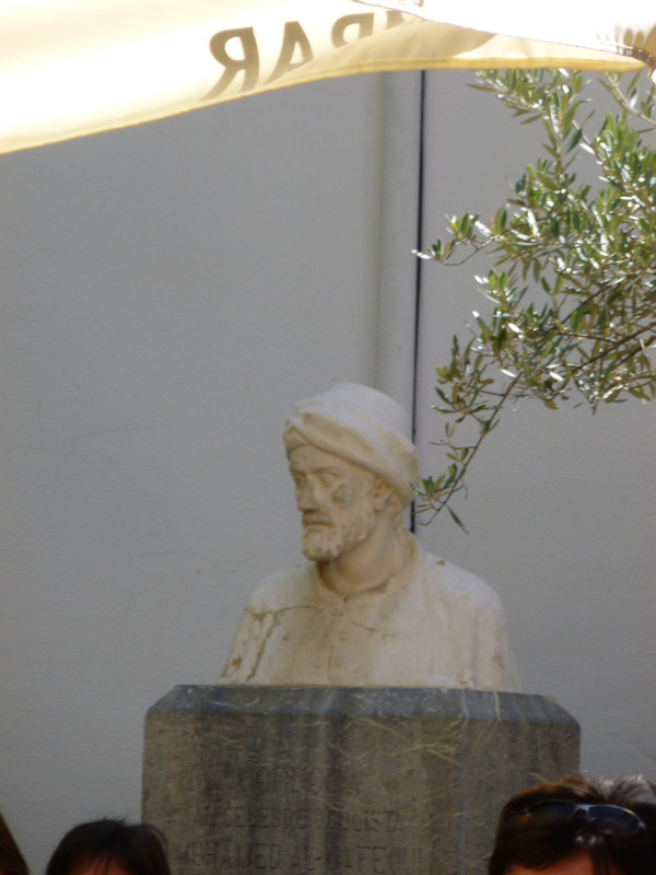 White stone bust on a grey-green stone plinth. Al-Ghafiqi is also wearing a cloth cap and a robe.