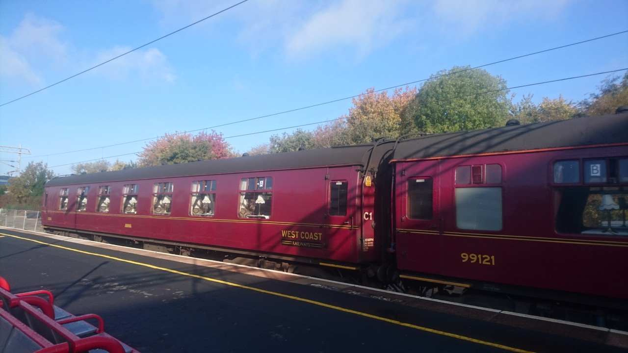 Two maroon red train carriages. The train carriage in the back has West Coast Railways written on it in gold. The front carriage has the number 99121 written on it in gold. Through the windows you can see that each table has a table lamp on it. That is a fancy train.