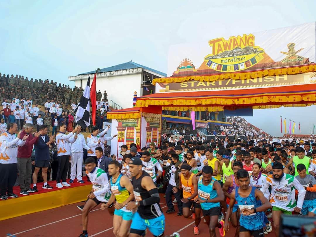 A male runner wearing a yellow singlet and blue shorts crosses a red-carpeted race finish area with his arms outstretched.