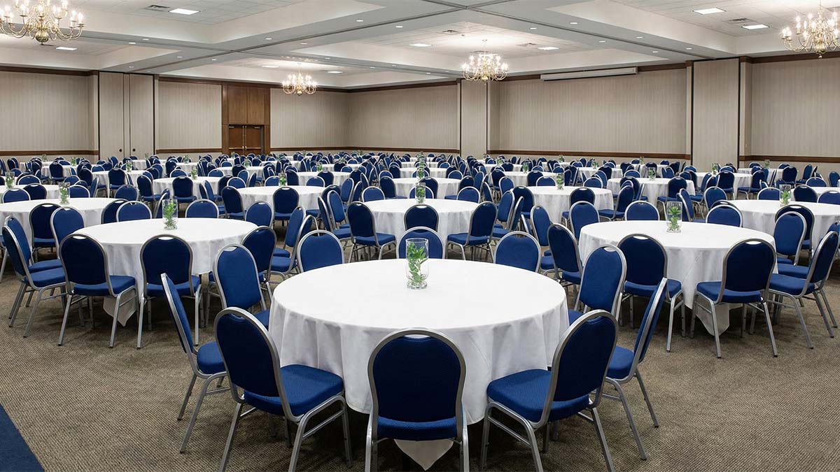 Большой банкетный зал с множеством одинаковых синих стульев с мягкой обивкой, расставленных вокруг круглых столов.