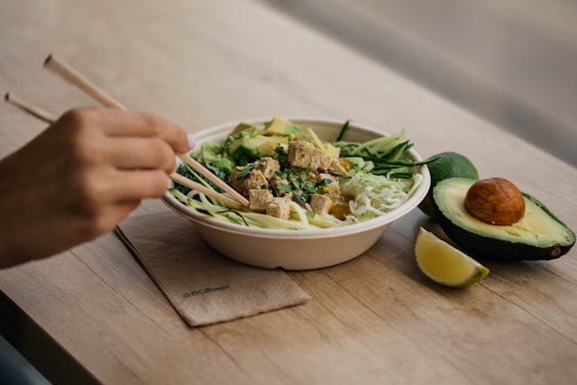 Woman hand holding chopsticks placed in avocado salad bowl, with half cut avocado and lemon slice placed beside the bowl on a table.