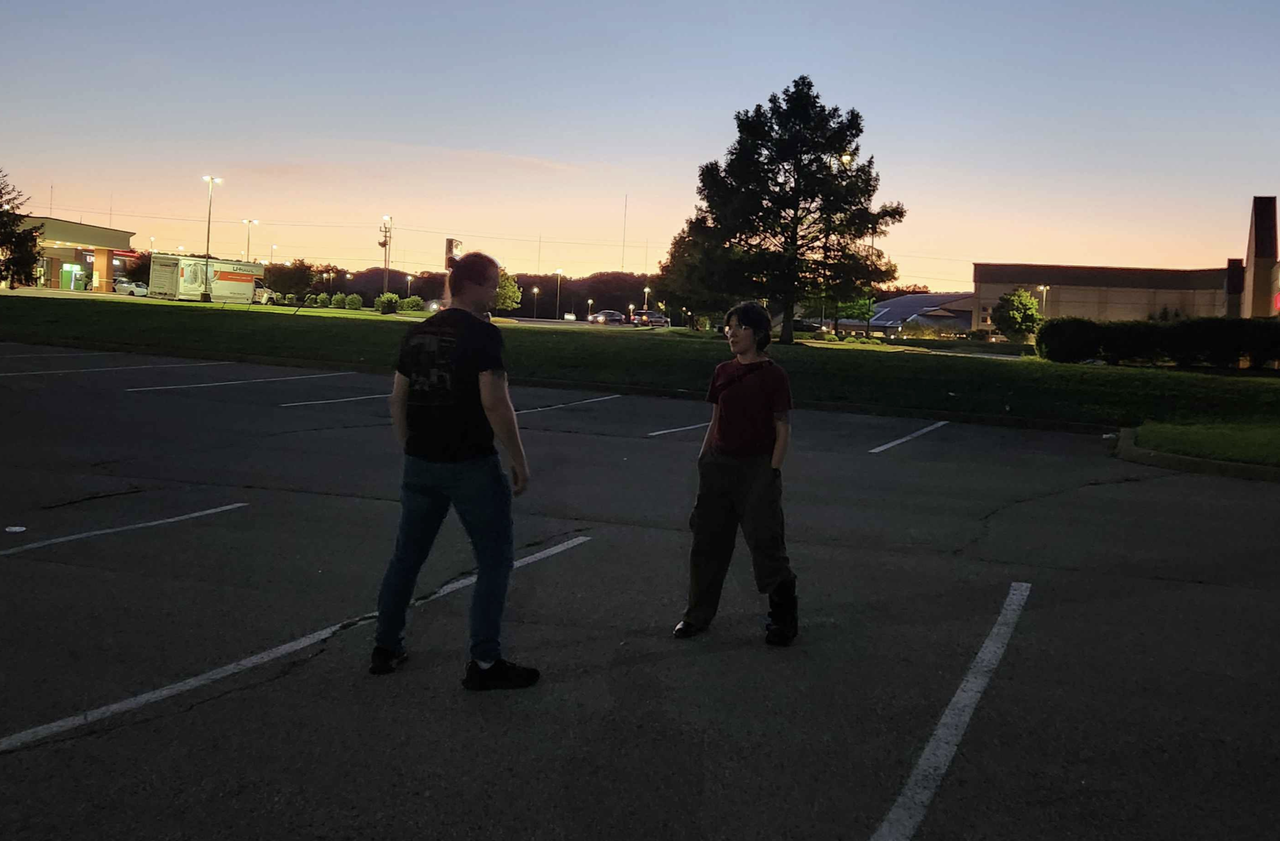 Mel and Seth facing off in a parking lot at sunset