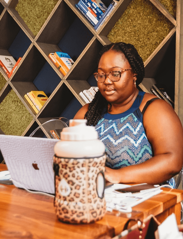Woman concentrating on her laptop in a coworking space with a stylish bookshelf behind her.