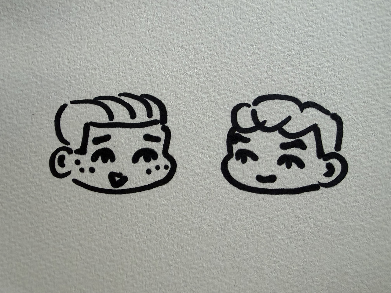 A drawing of two small heads using a black marker. The head on the left has swept back hair with freckles, looking happy. The head on the right has a bit fluffy, swept back hair too, smiling at the other one.