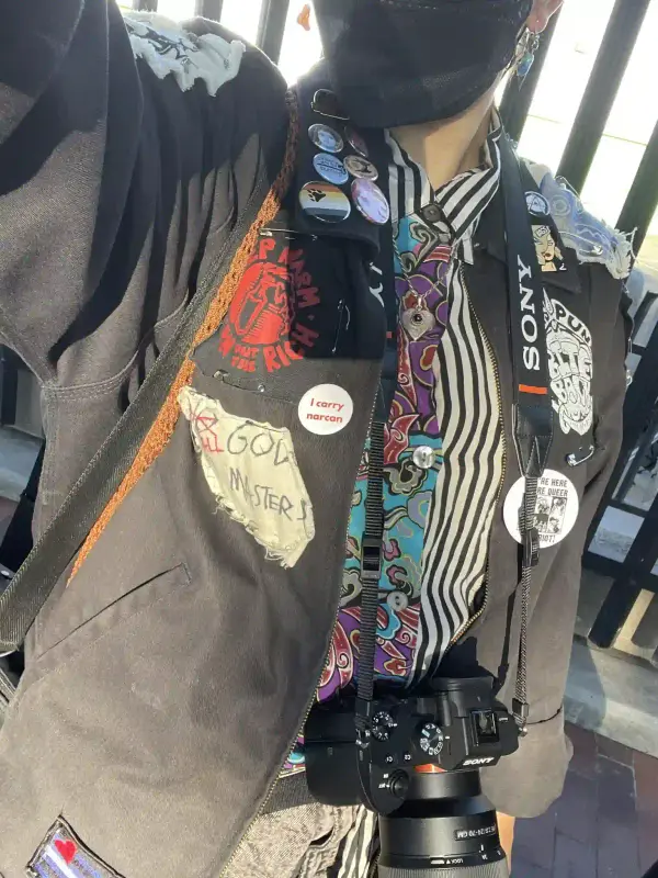 a torso selfie of myself with my battle jacket and a fun printed button up underneath. you can see a DSLR hanging around my neck and the strap of a bag on my shoulder.