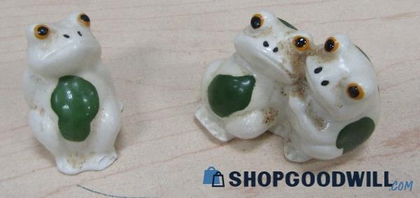 a product photo of two small white ceramic frog figurines. on the left is a grumpy-looking frog with a blob of green on its belly. on the right is one statue of two frogs; one is leaning on the other, who is gripping its shoulder supportively.