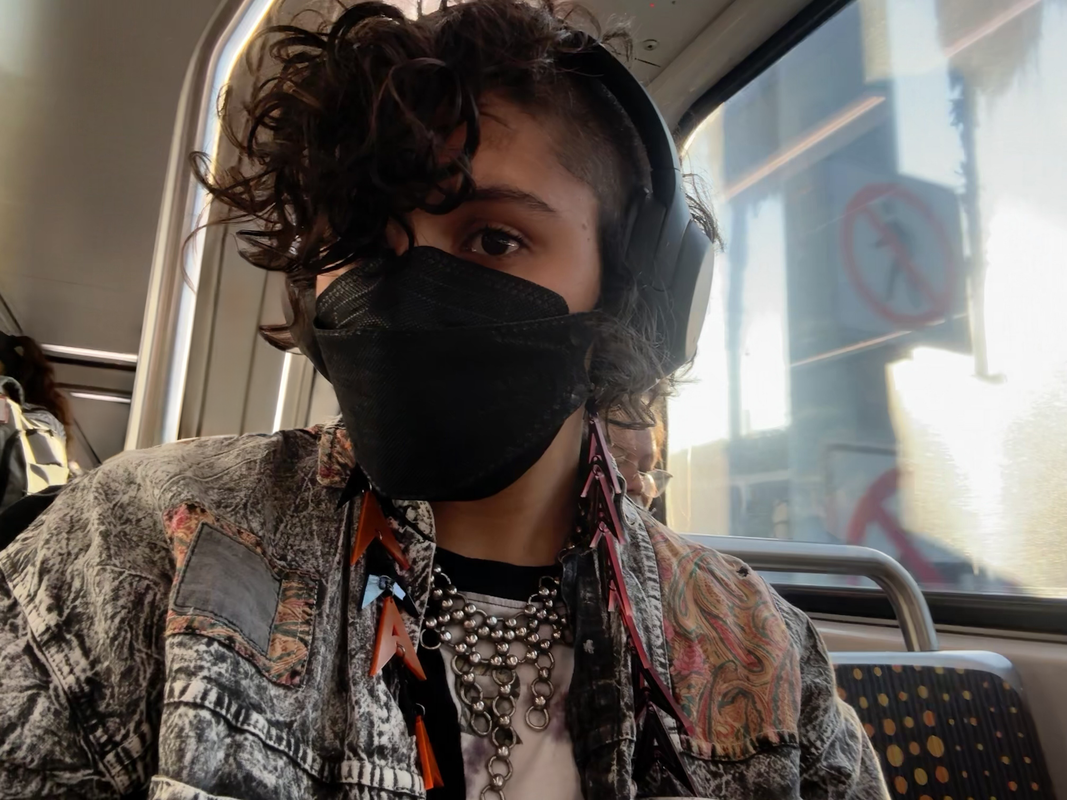 a selfie of a person with light brown skin and a mohawk on a train. zie is wearing black over-ear headphones, a metal necklace, an overshirt, and long earrings.