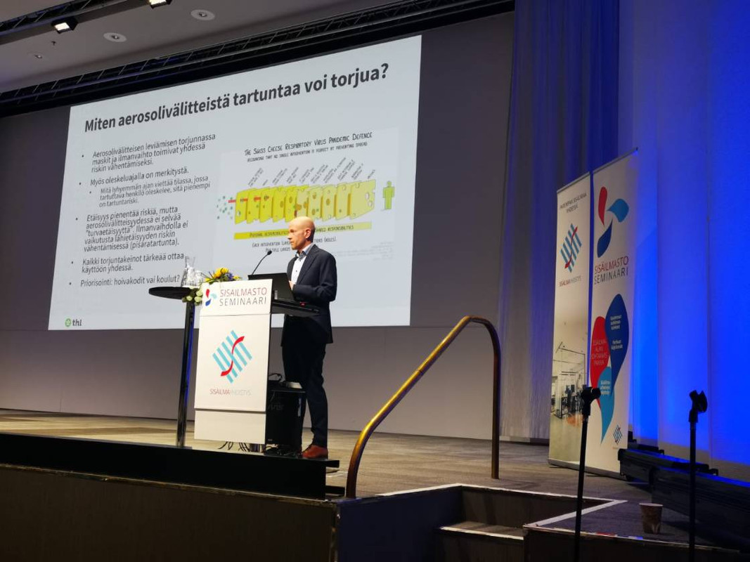 THL:n Terveysturvaajat-osaston johtava ylilääkäri Otto Helve puhumassa Sisäilmayhdistyksen seminaarissa korona ilmalevitteisyydstä. Taustalle heijastettuna näkyy "reikäjuustomalli" 