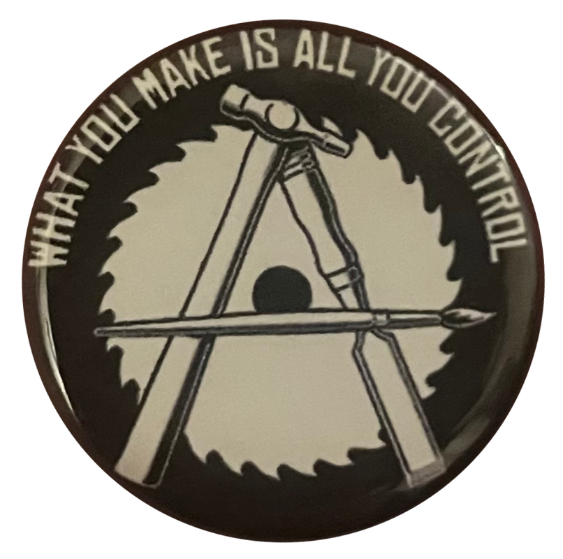a black pin with white text/drawings that says 'what you make is all you control' and an anarchist 'A' symbol crafted from a hammer, chisel, and paint brush, all in front of a buzz saw in back